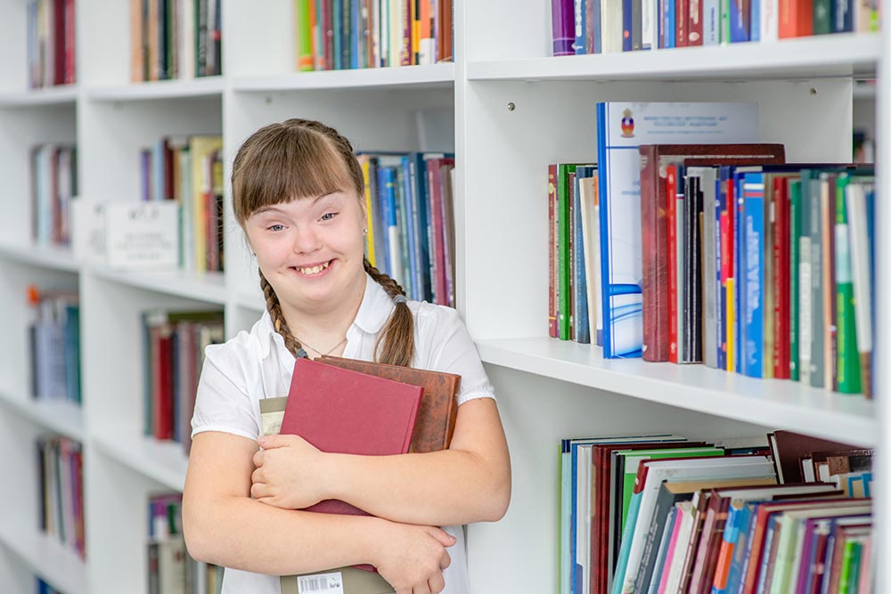 Young female student with Downs Syndrome in a library holding books and smiling.
